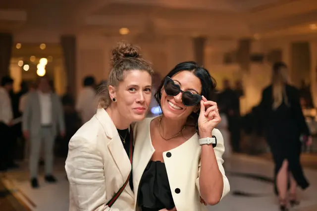 Two women in cream blazers, one lifting heart-shaped sunglasses
